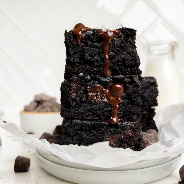 vegan brownies stacked on a white plate next to a bottle of milk and a bowl of chocolate chips