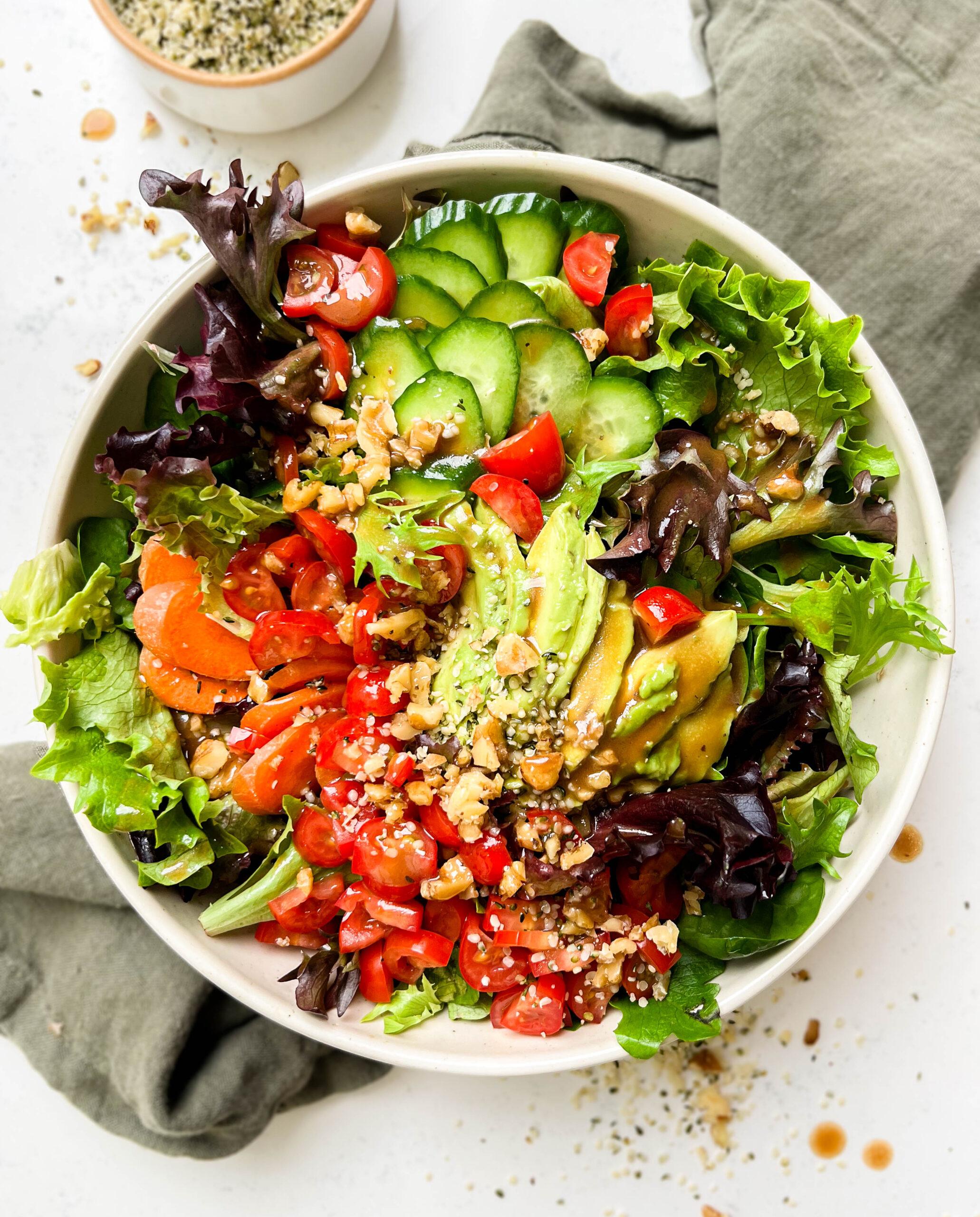 classic garden salad with tomatoes, carrots, avocado, greens and nuts in a bowl on a green linen cloth with balsamic dressing on top