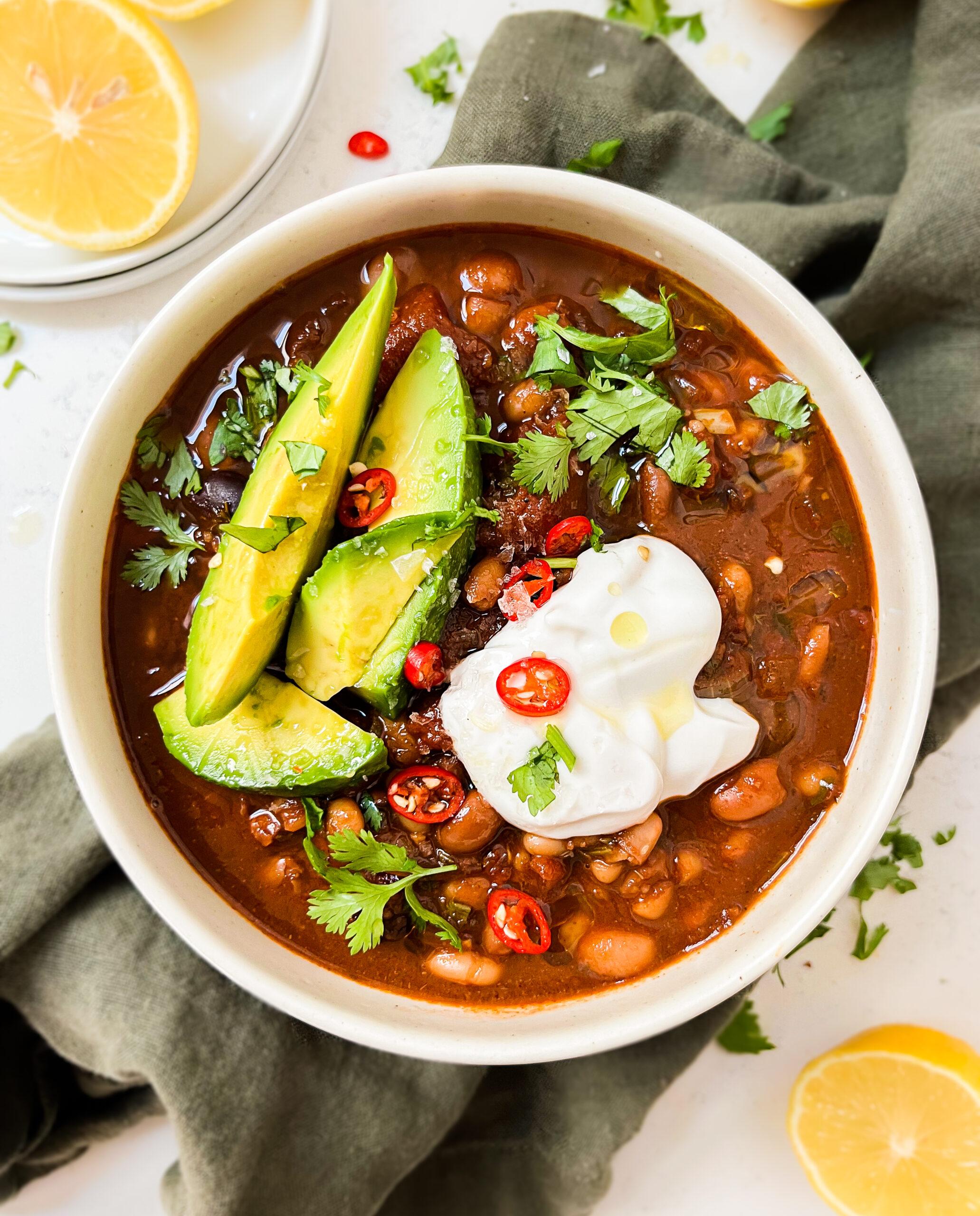 chili in a bowl with avocado, yogurt and spices on top next to a green cloth and lemons