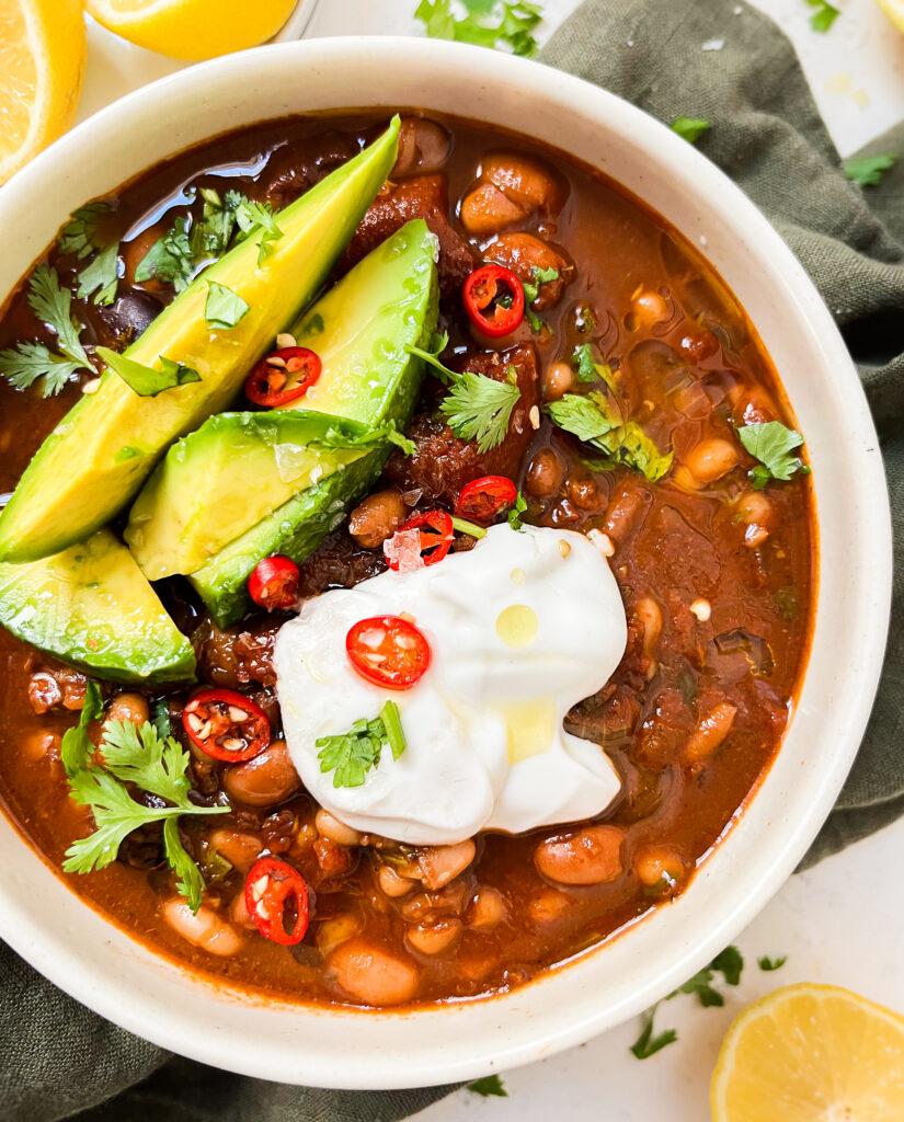 chili in a bowl with avocado, yogurt and spices on top next to a green cloth and lemons