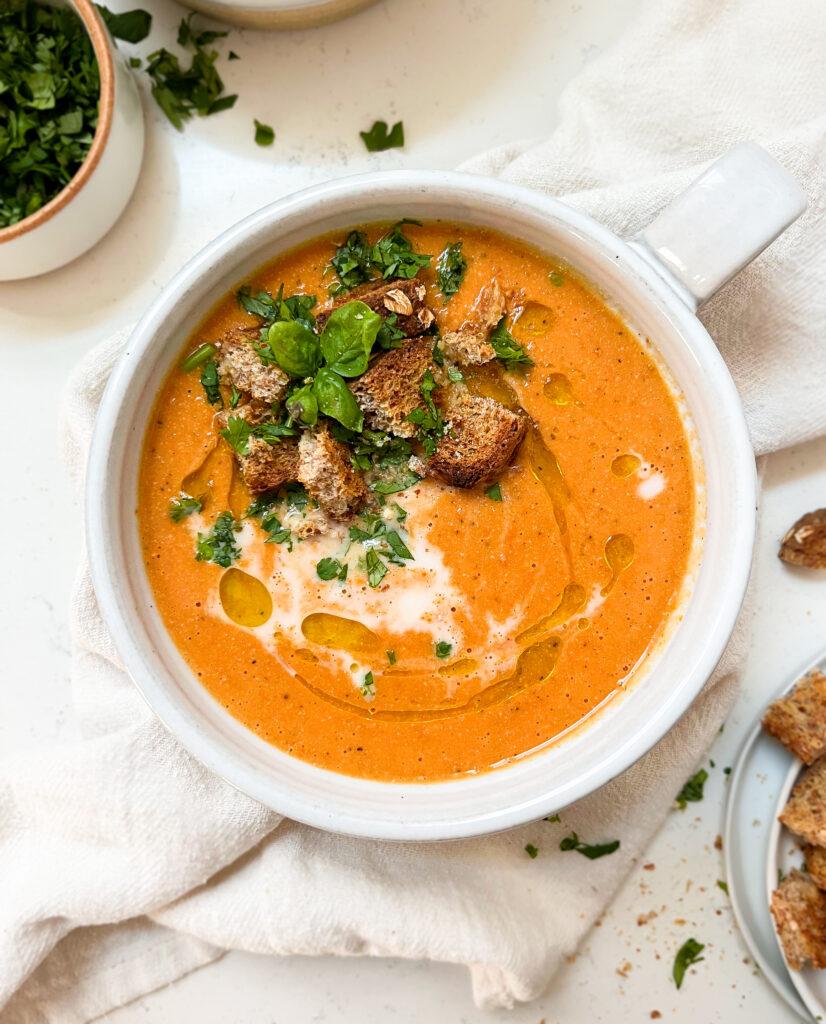 hands holding a white soup with vegan roasted tomato soup inside with croutons and basil on top