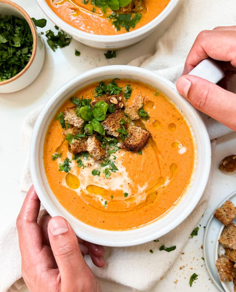 hands holding a white soup with vegan roasted tomato soup inside with croutons and basil on top