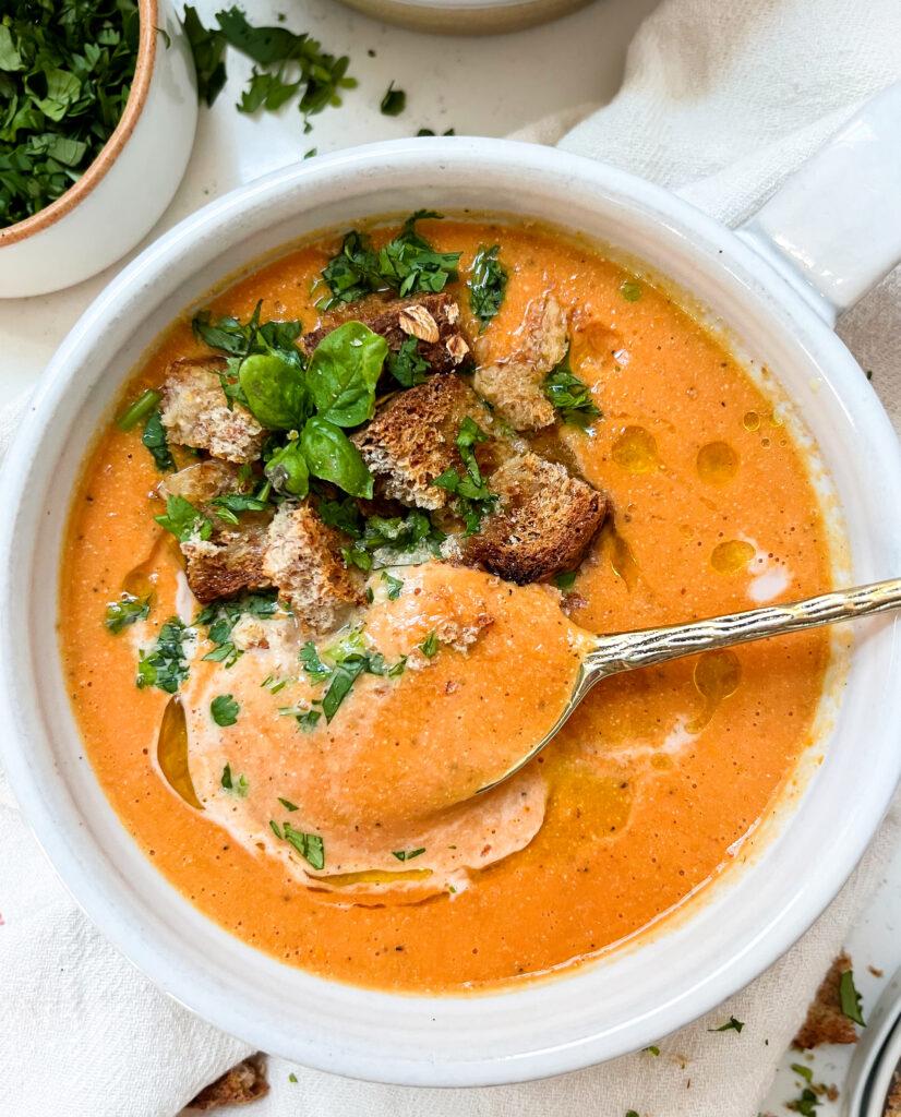 gold spoon in a white soup with vegan roasted tomato soup inside with croutons and basil on top