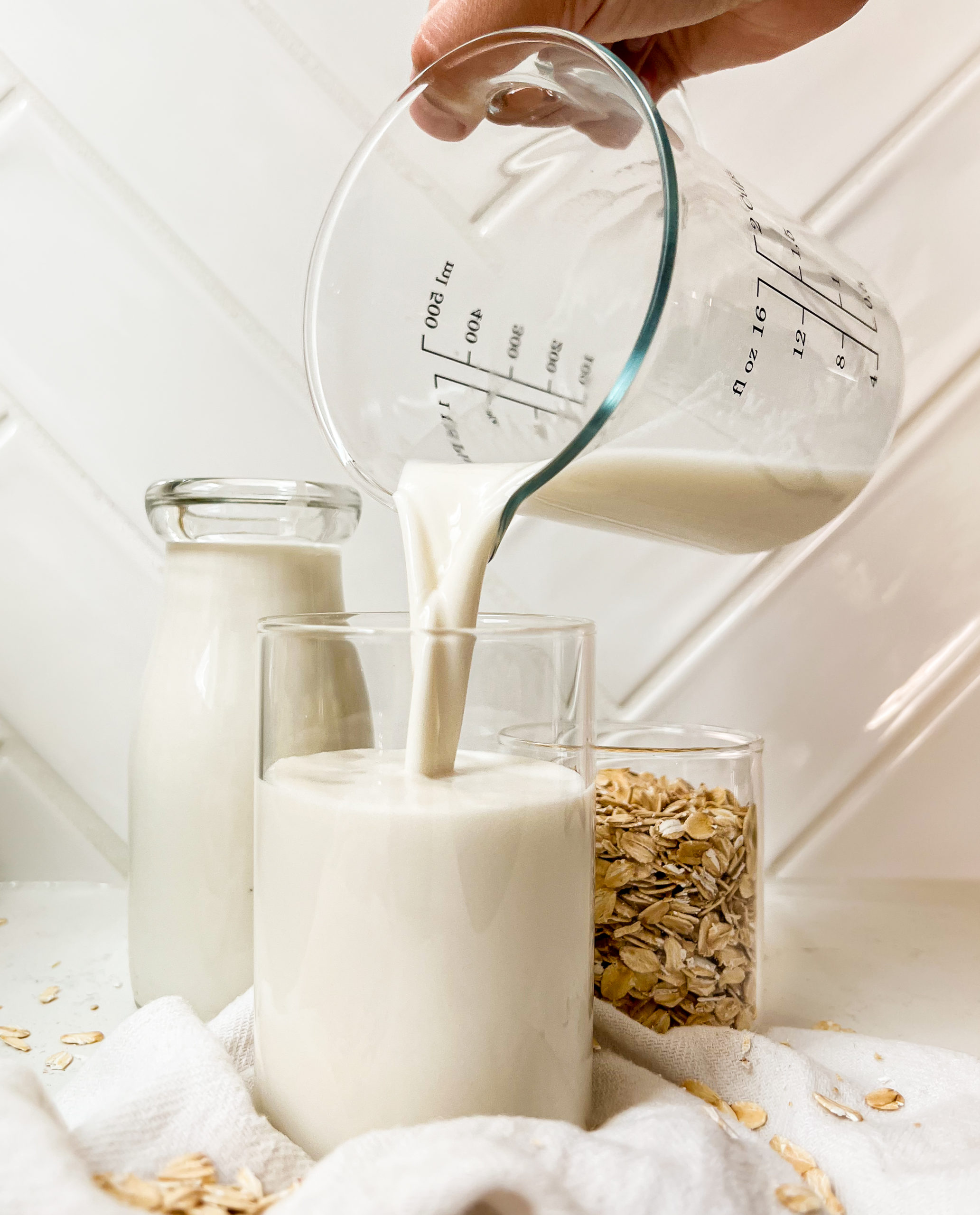oat milk pouring into a cup next to a jar of milk and a cup of rolled oats