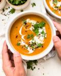 hands holding a bowl of creamy red lentil soup next to a bowl of cilantro