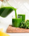 pitcher pouring green juice into a glass on wooden cutting board next to kale and lemons