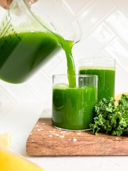 pitcher pouring green juice into a glass on wooden cutting board next to kale and lemons