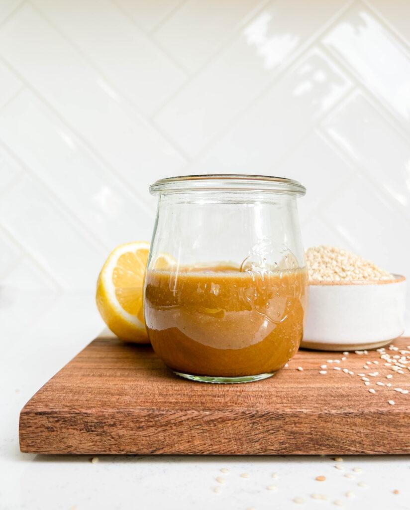 peanut dressing in a glass jar on a cutting board next to lemons and a bowl of sesame seeds