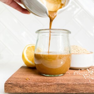 peanut dressing in a glass jar on a cutting board next to lemons and a bowl of sesame seeds