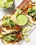 spicy tempeh tacos with a creamy jalapeno verde sauce on a cutting board next to a bowl of lettuce and limes