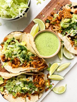 spicy tempeh tacos with a creamy jalapeno verde sauce on a cutting board next to a bowl of lettuce and limes