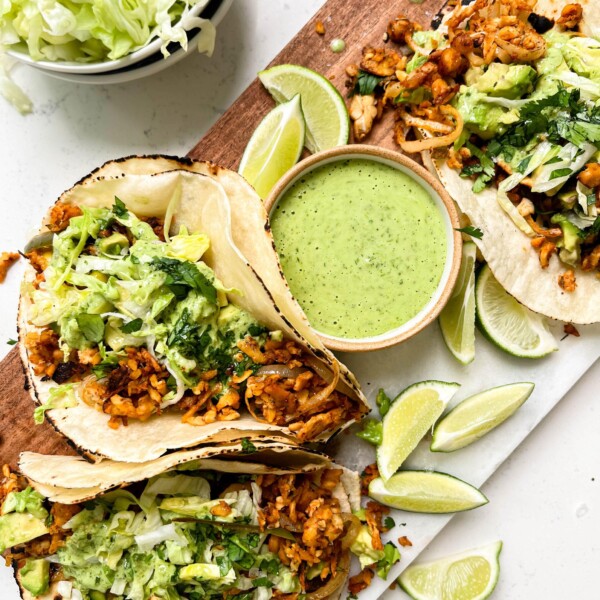 spicy tempeh tacos with a creamy jalapeno verde sauce on a cutting board next to a bowl of lettuce and limes