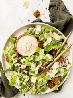 a beige plate with vegan caesar salad, two gold forks and a bowl of dressing on top