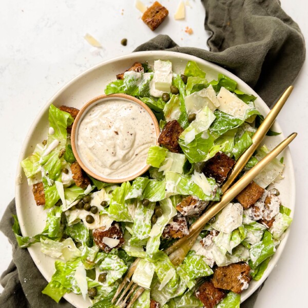 a beige plate with vegan caesar salad, two gold forks and a bowl of dressing on top