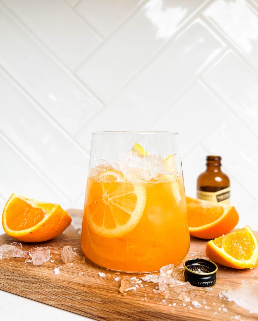 a glass of orange spritz on a wooden cutting board next to sliced oranges and vanilla extract
