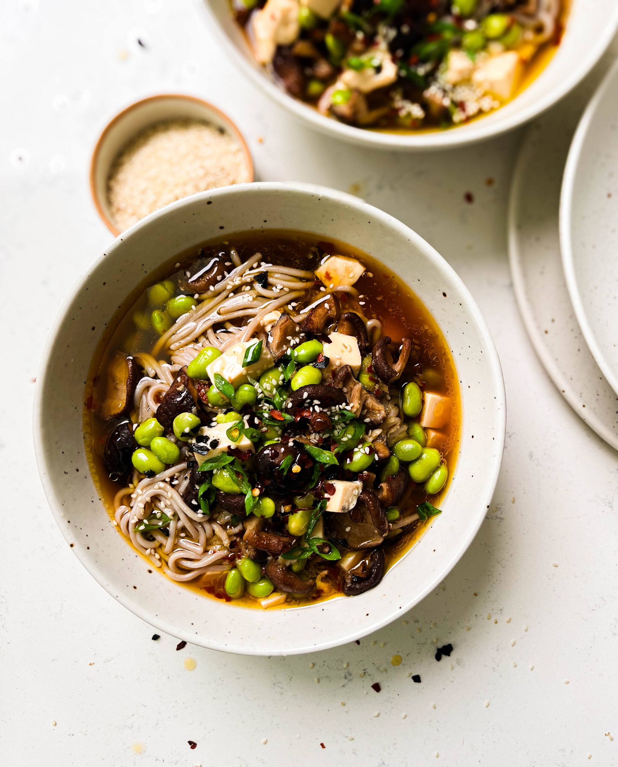 two bowls of spicy mushroom noodle soup next to a bowl of sesame seeds