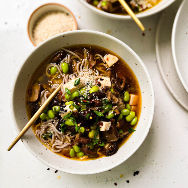 two bowls of spicy mushroom noodle soup next to a bowl of sesame seeds