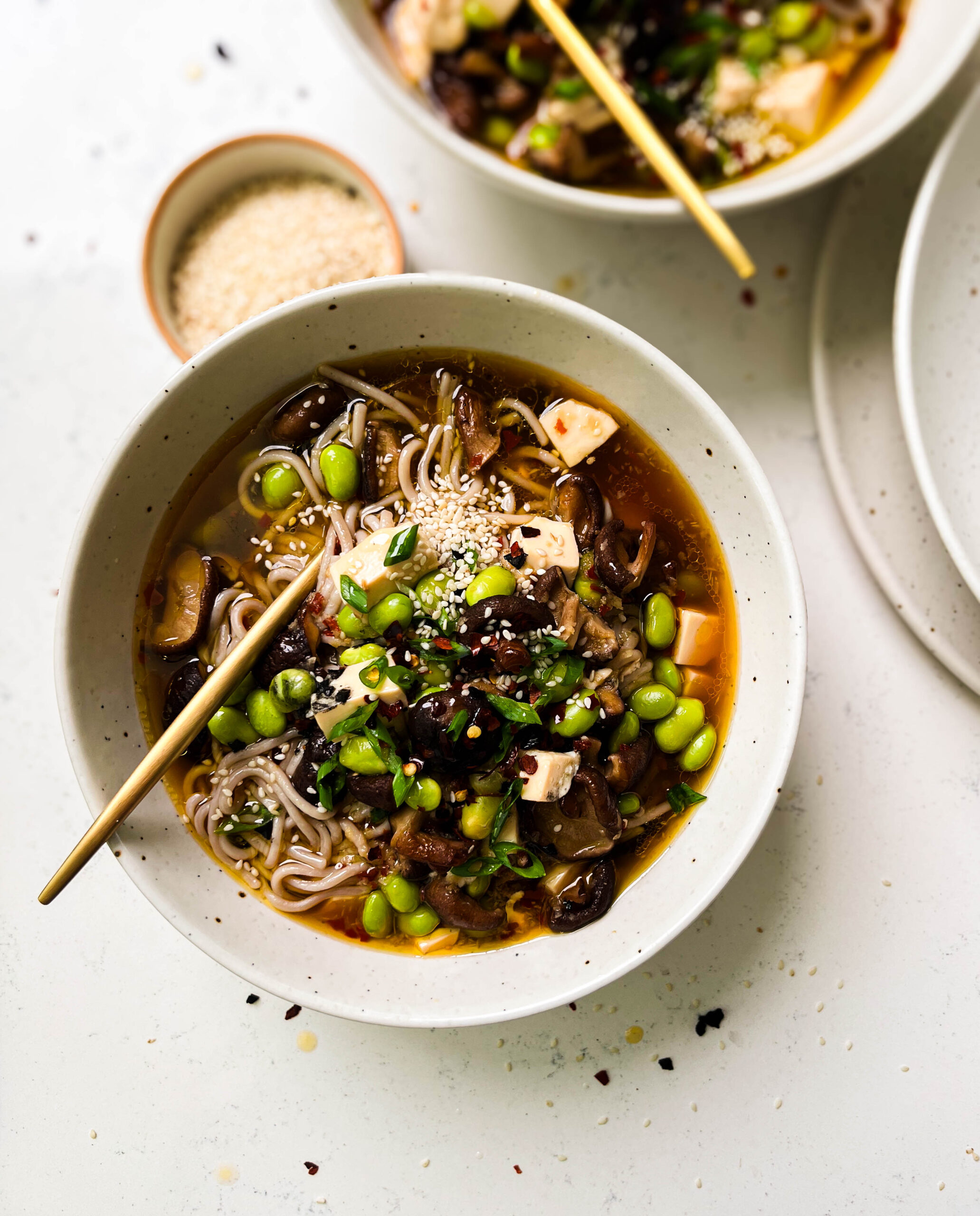 two bowls of spicy mushroom noodle soup next to a bowl of sesame seeds
