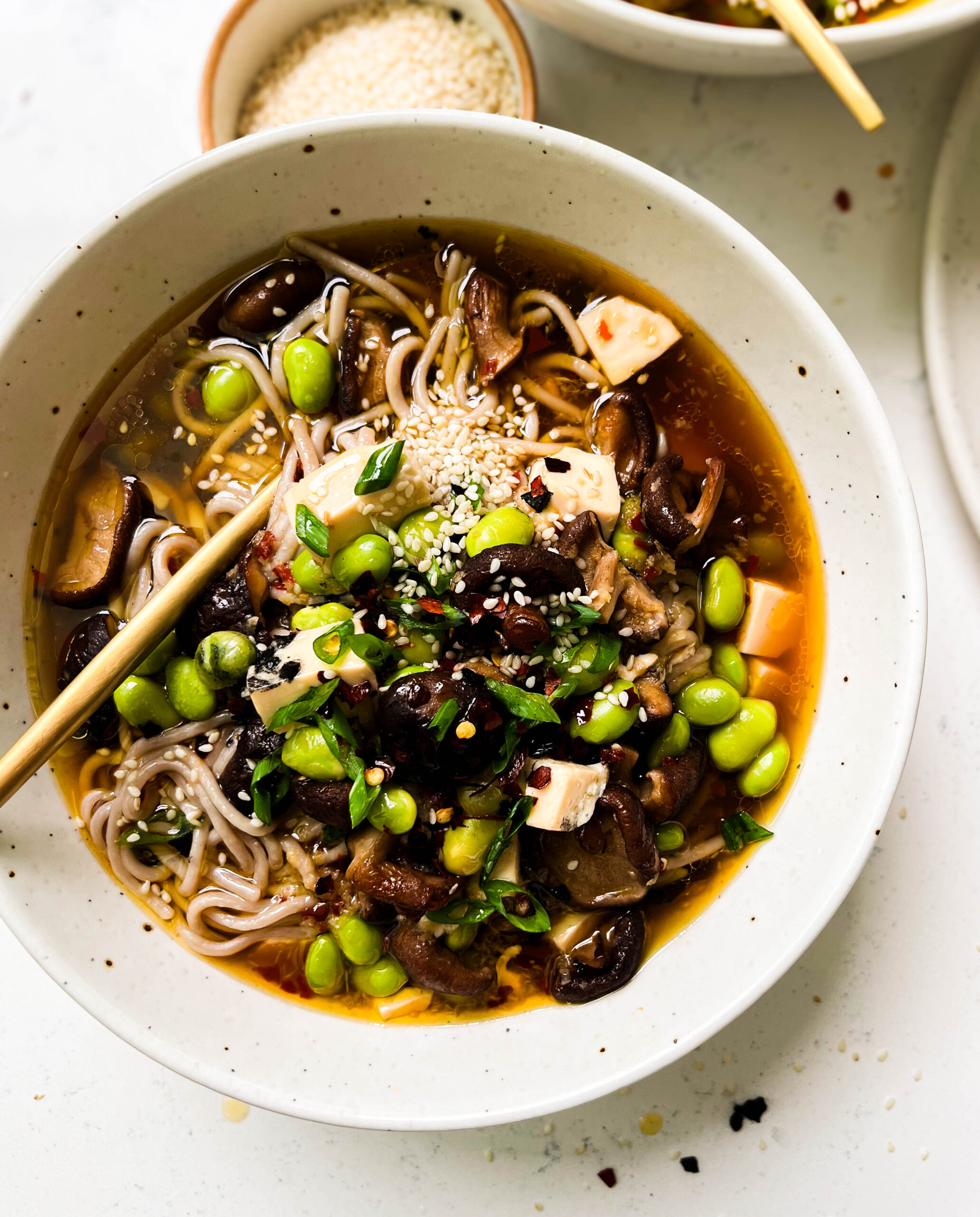 bowl of spicy mushroom noodle soup next to a bowl of sesame seeds